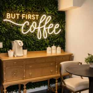 But First Coffee LED neon sign in warm white and gold mounted on a green plant wall above a wooden coffee bar console
